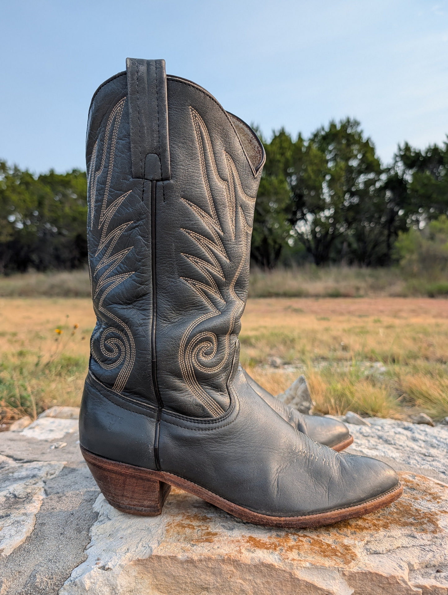 A pair of dark gray leather cowboy boots with decorative light-tan stitching, resting on a stone surface outdoors with a grassy field and trees in the background.