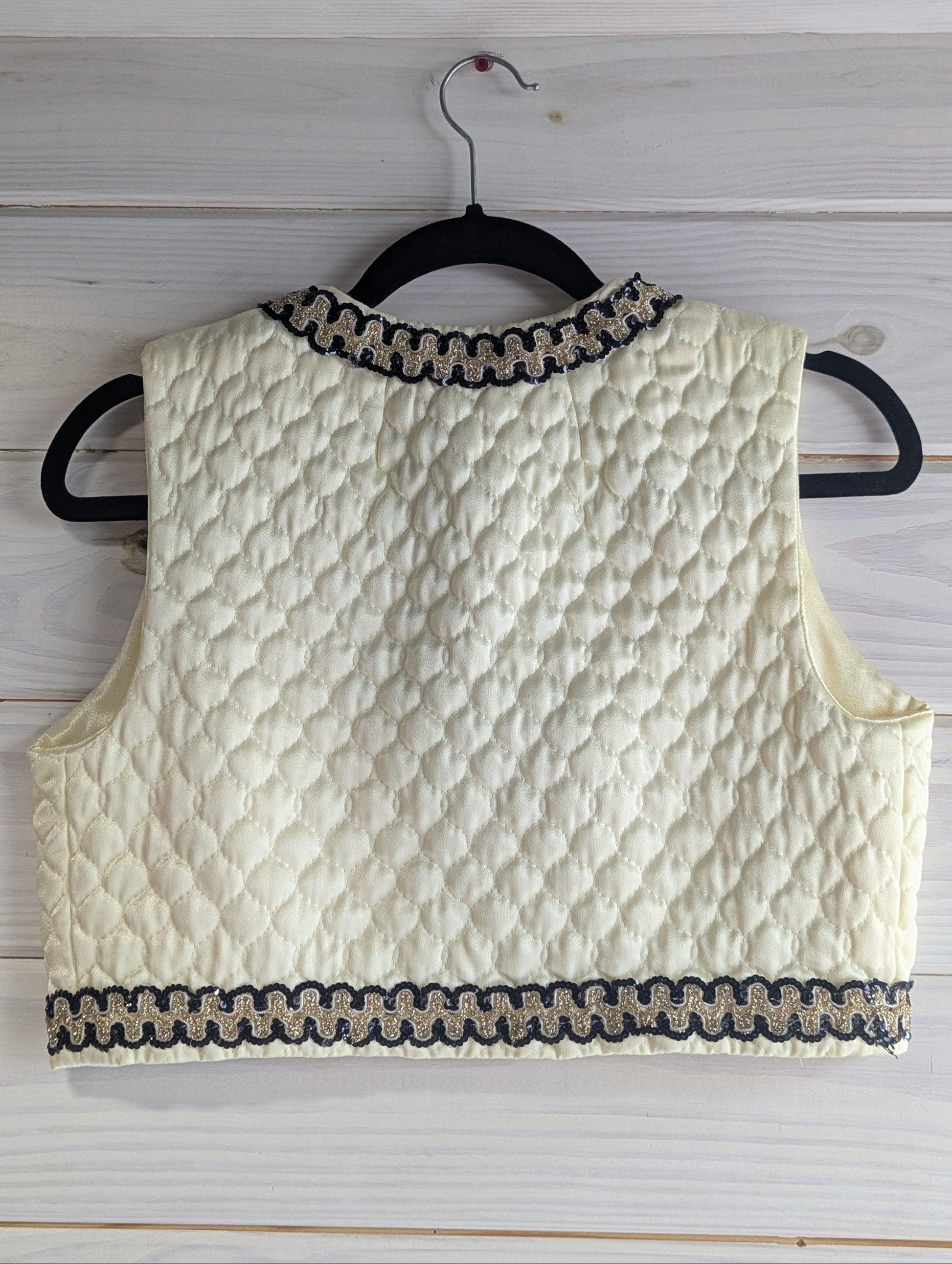 Ivory quilted vest with black and gold sequin trim on a hanger against a wooden background