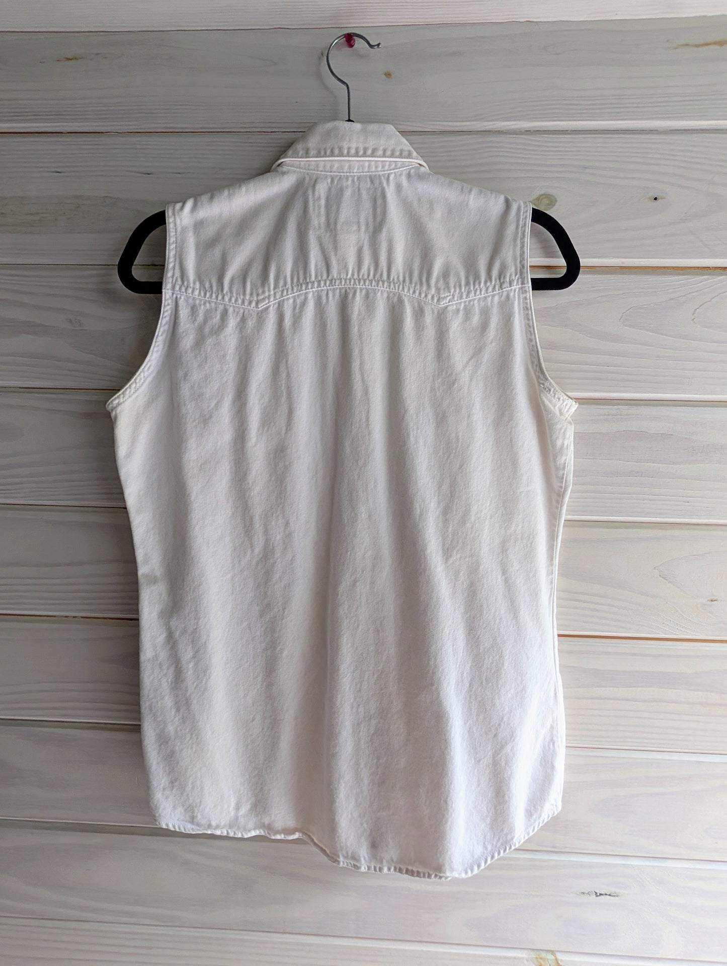 Back of white sleeveless 100% cotton denim shirt with white and silver pearl snap buttons on a hanger against a wooden background. Western style horizontal hemming on top of shoulders in slight double point on top of back. 