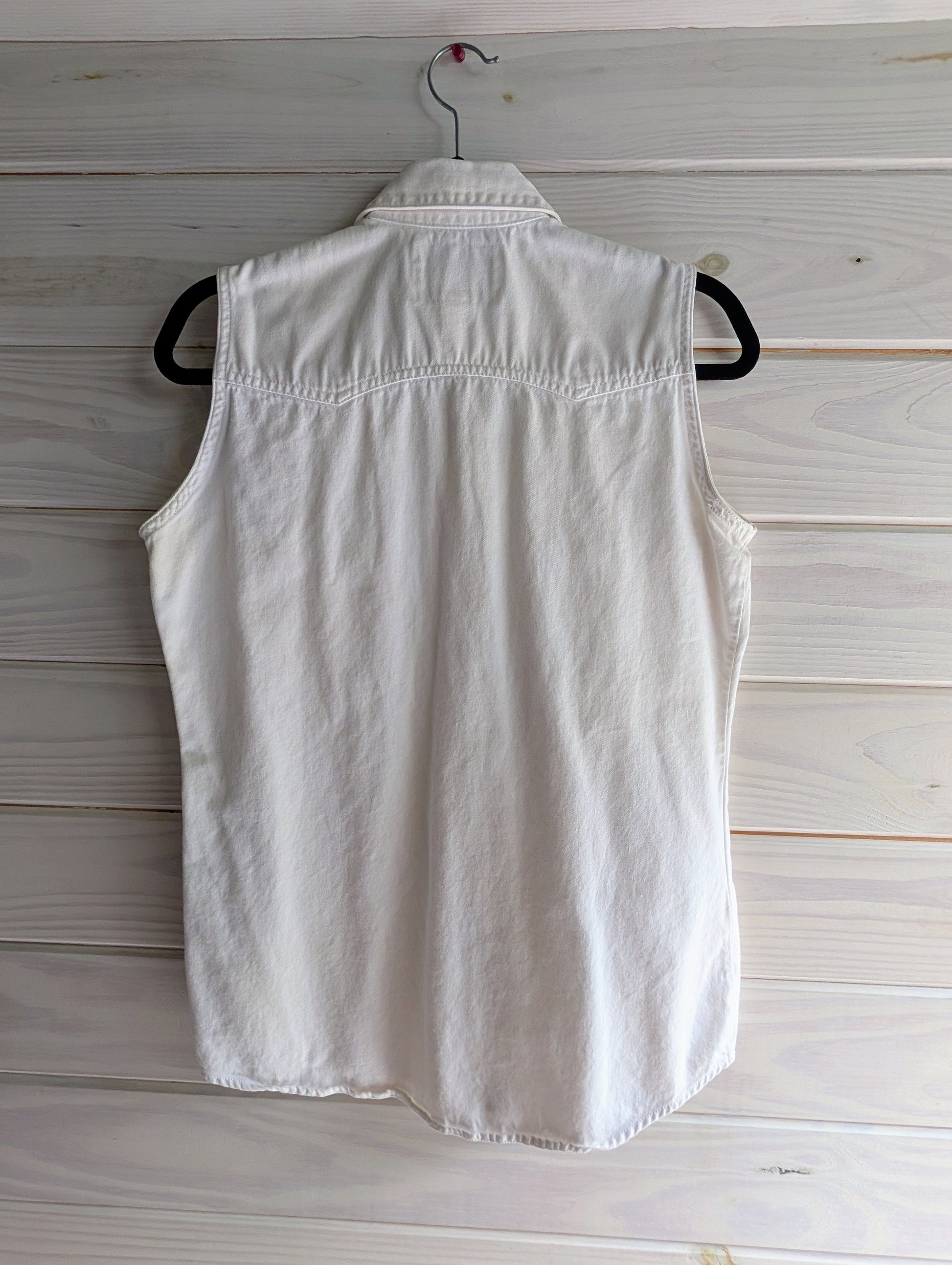 Back of white sleeveless 100% cotton denim shirt with white and silver pearl snap buttons on a hanger against a wooden background. Western style horizontal hemming on top of shoulders in slight double point on top of back. 