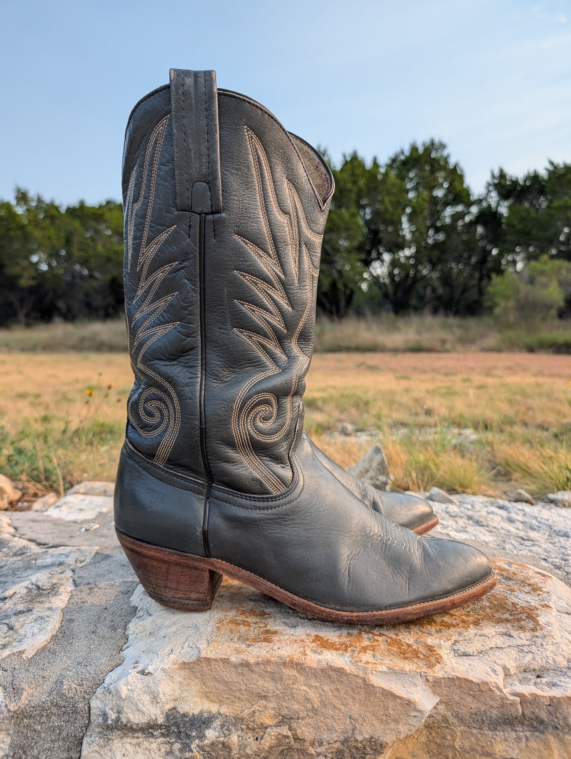 Dark gray leather cowboy boot with decorative stitching in cream thread, a 2" Western heel, resting on a large stone outdoors with a grassy field and trees in the background. 