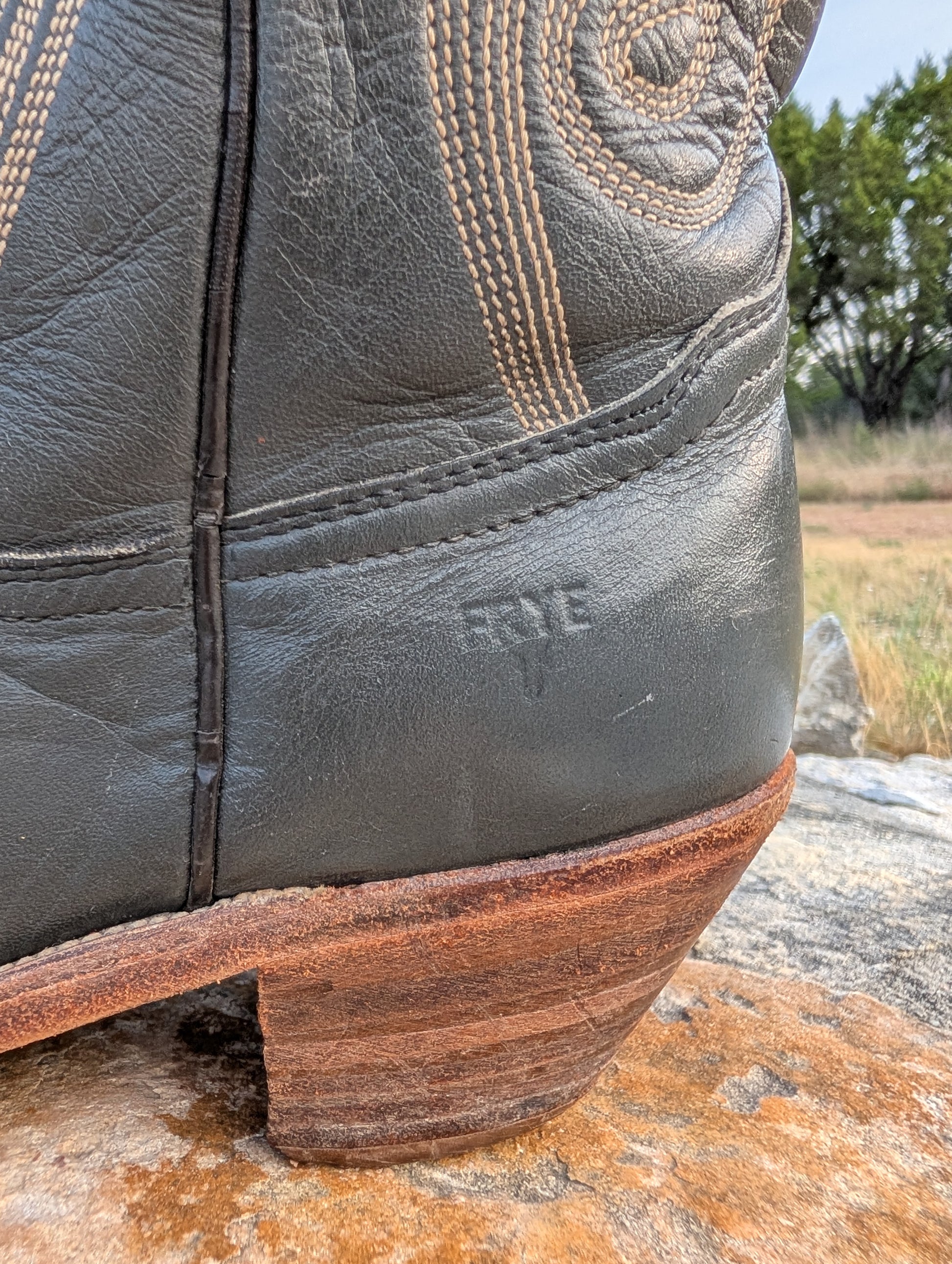 Close-up of a dark gray leather cowboy boot resting on a rock outdoors, showing the heel, sole edge, decorative stitching, and embossed FRYE logo on the leather above the heel. 