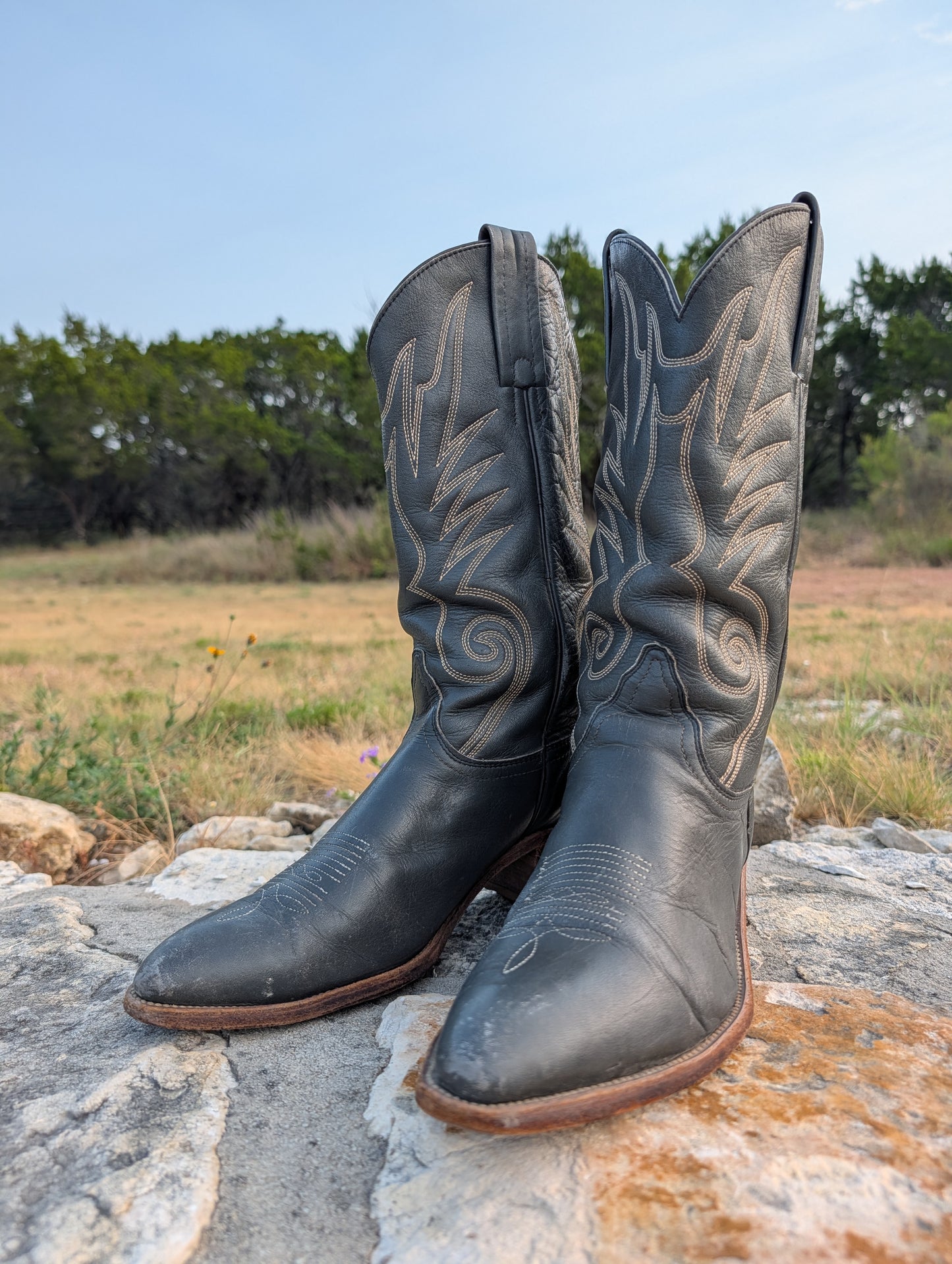 A pair of gray leather cowboy boots with light tan western embroidery, resting on a stone surface outdoors with a grassy field and trees in the background.