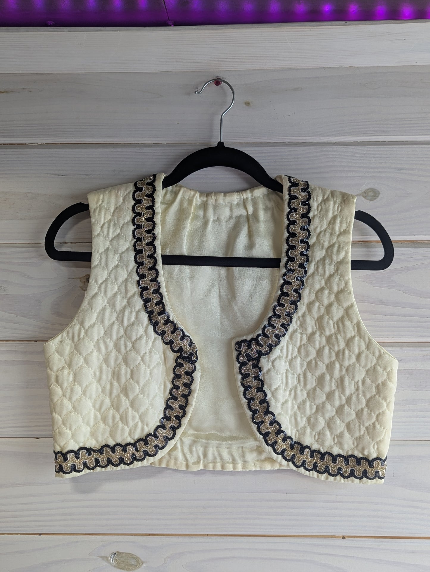 Ivory quilted vest with black and gold sequin trim on a hanger against a wooden background