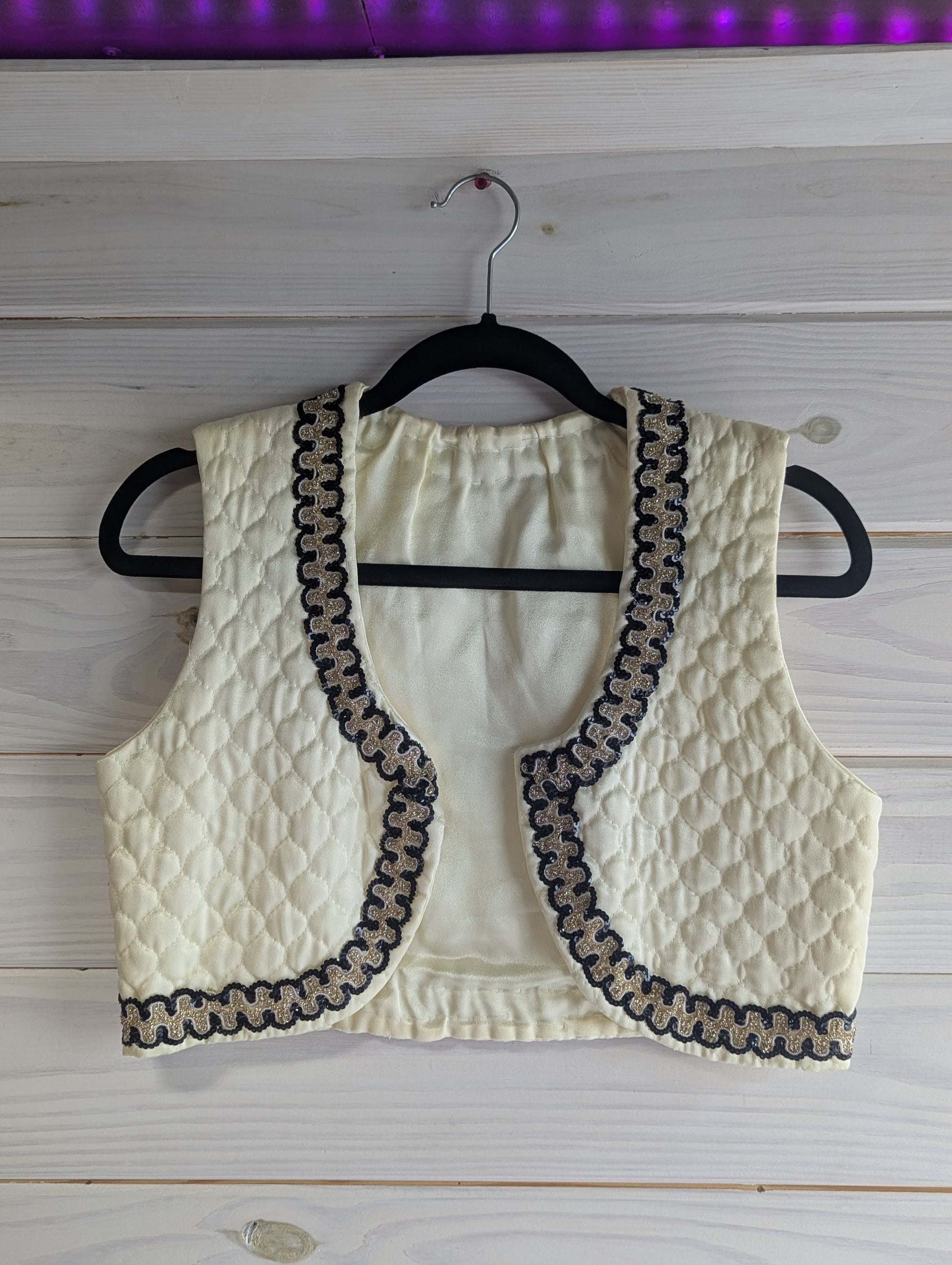 Ivory quilted vest with black and gold sequin trim on a hanger against a wooden background