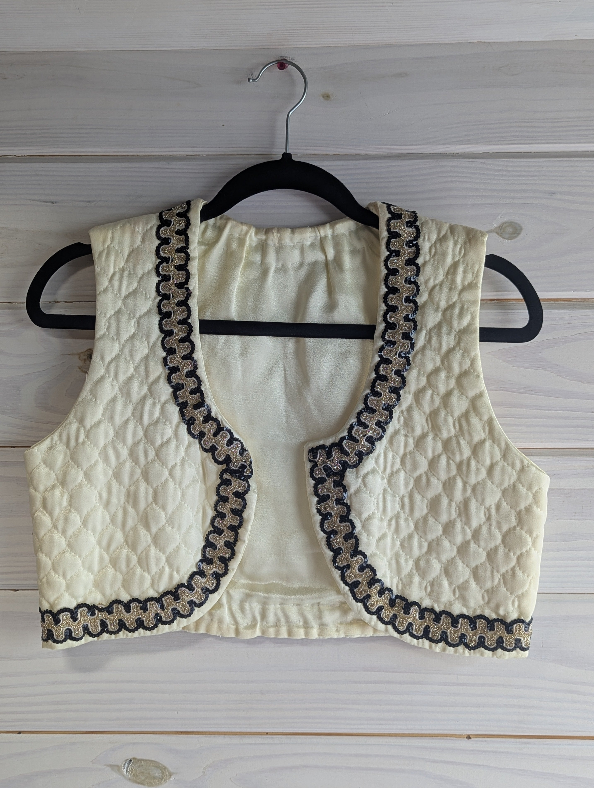 Ivory quilted vest with black and gold sequin trim on a wooden hanger against a wooden background