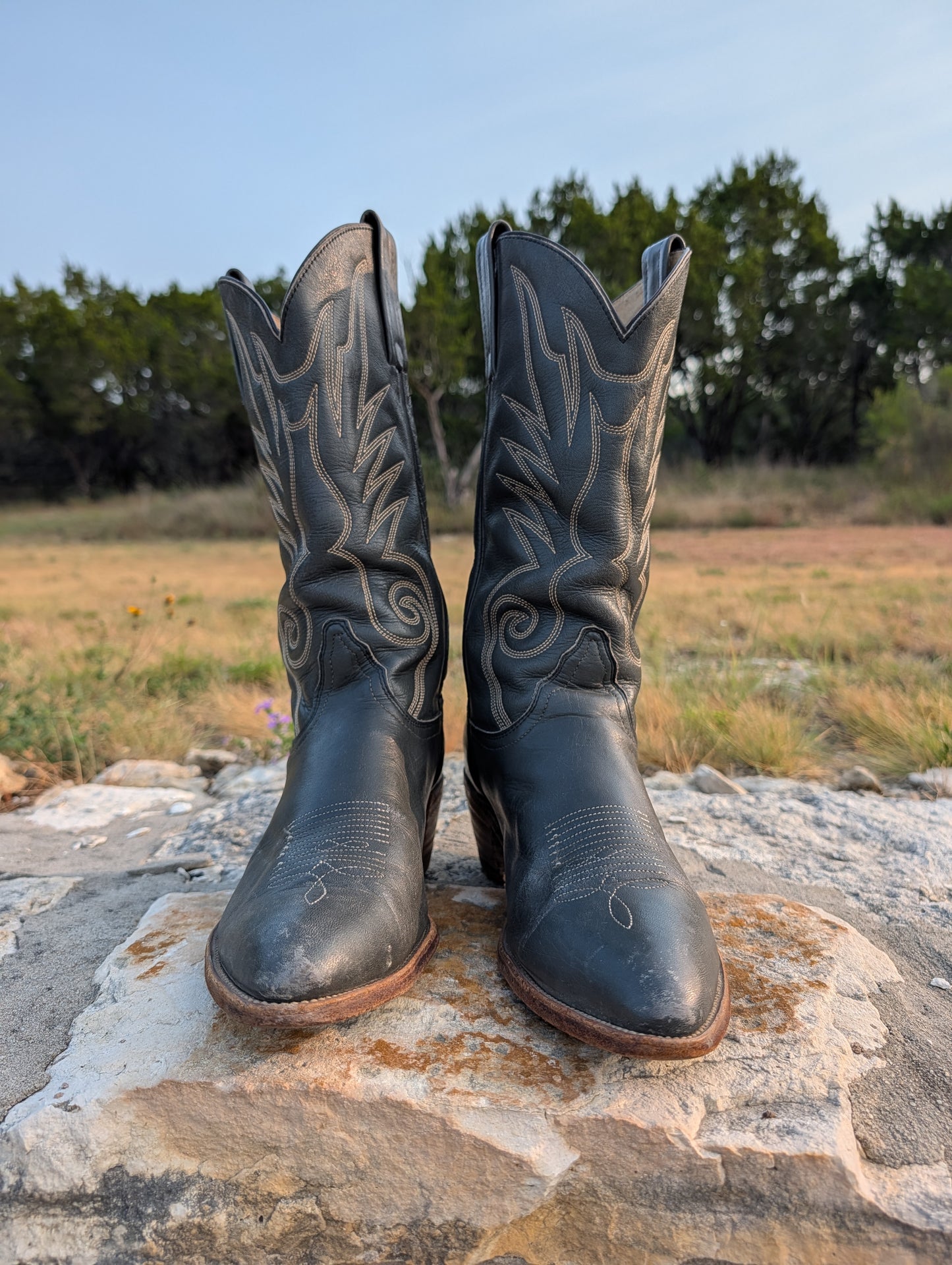 Dark gray leather cowboy boots facing forward with decorative stitching in cream thread, a 2" Western heel, resting on a large stone outdoors with a grassy field and trees in the background. 