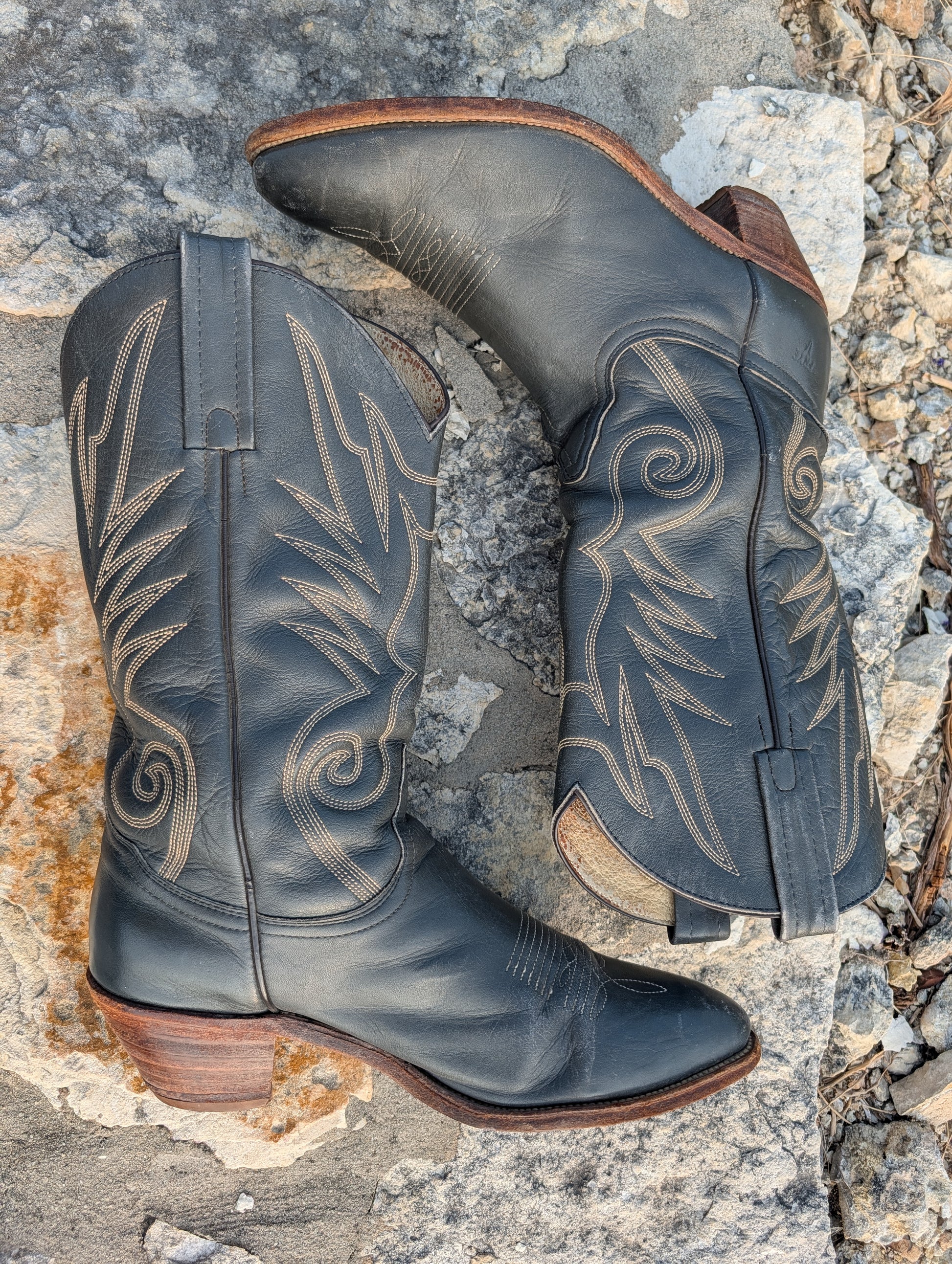 A pair of dark gray leather cowboy boots with tan western embroidery, displayed on a textured gray and brown rock surface, laying side by side.
