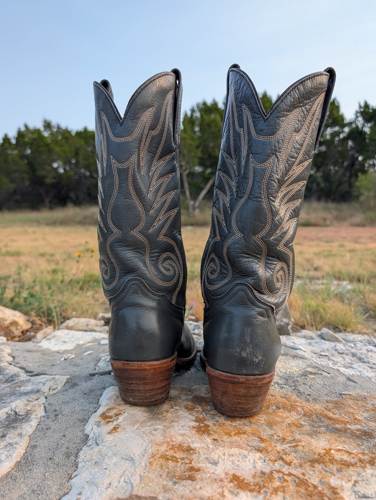 A pair of dark gray leather cowboy boots with western embroidery and brown heels, shown from the back while standing on a stone surface outdoors with a grassy field and trees in the background. 