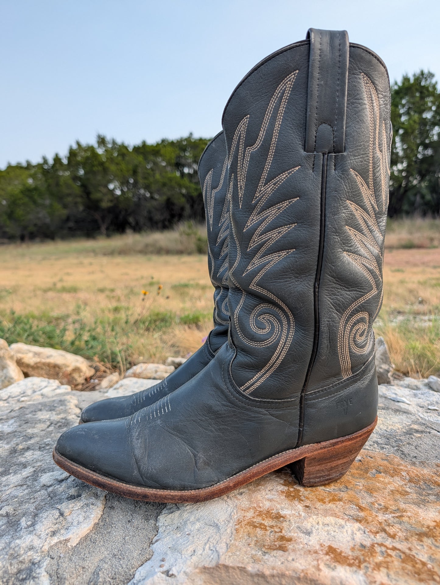 A pair of dark gray leather cowboy boots with light tan western embroidery, resting on a large rock in an outdoor grassy field.