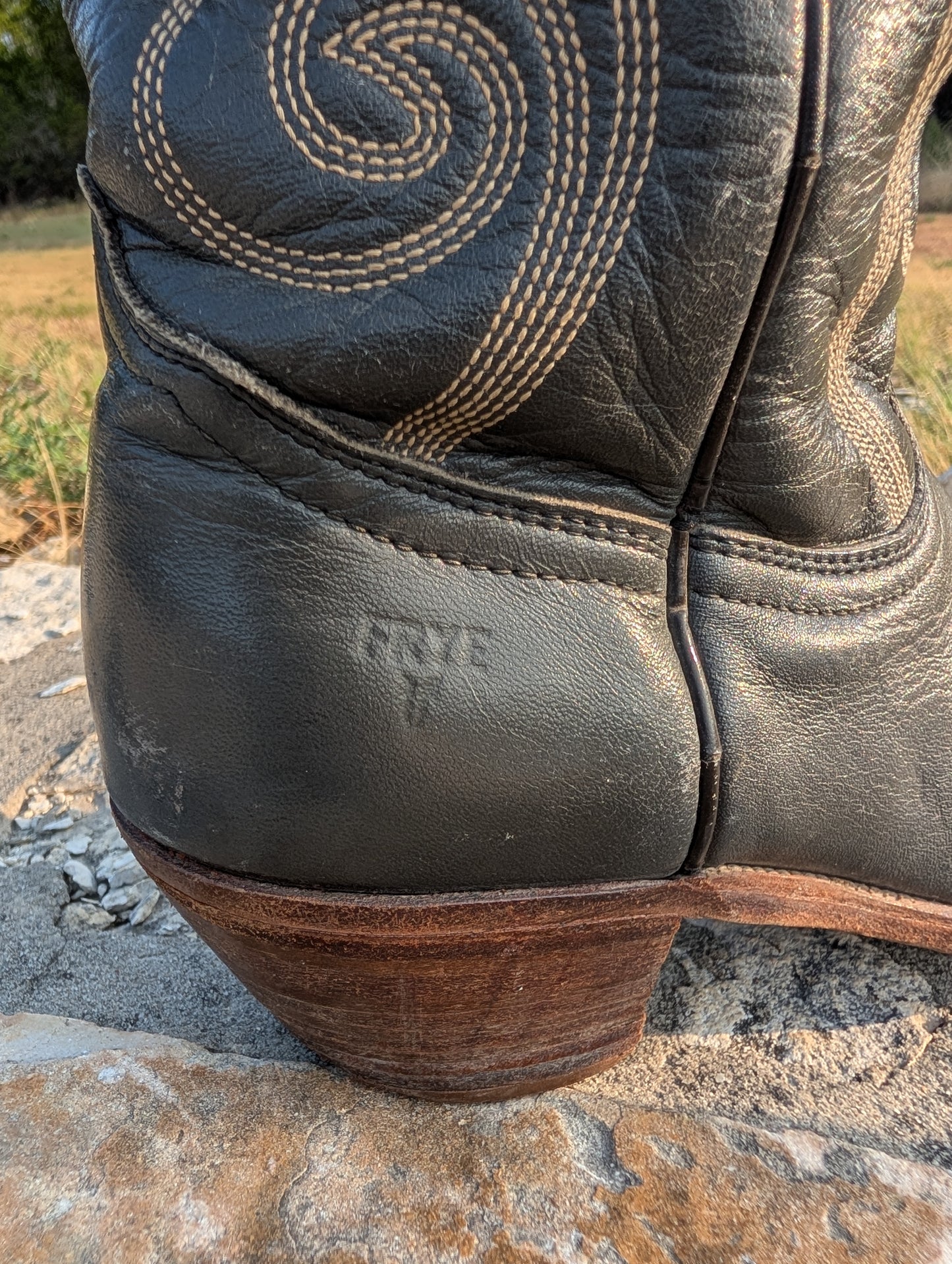 Close-up of a dark gray leather cowboy boot heel resting on a stone surface outdoors. The boot shows decorative light stitching on the shaft and an embossed 'FRYE' logo near the heel.
