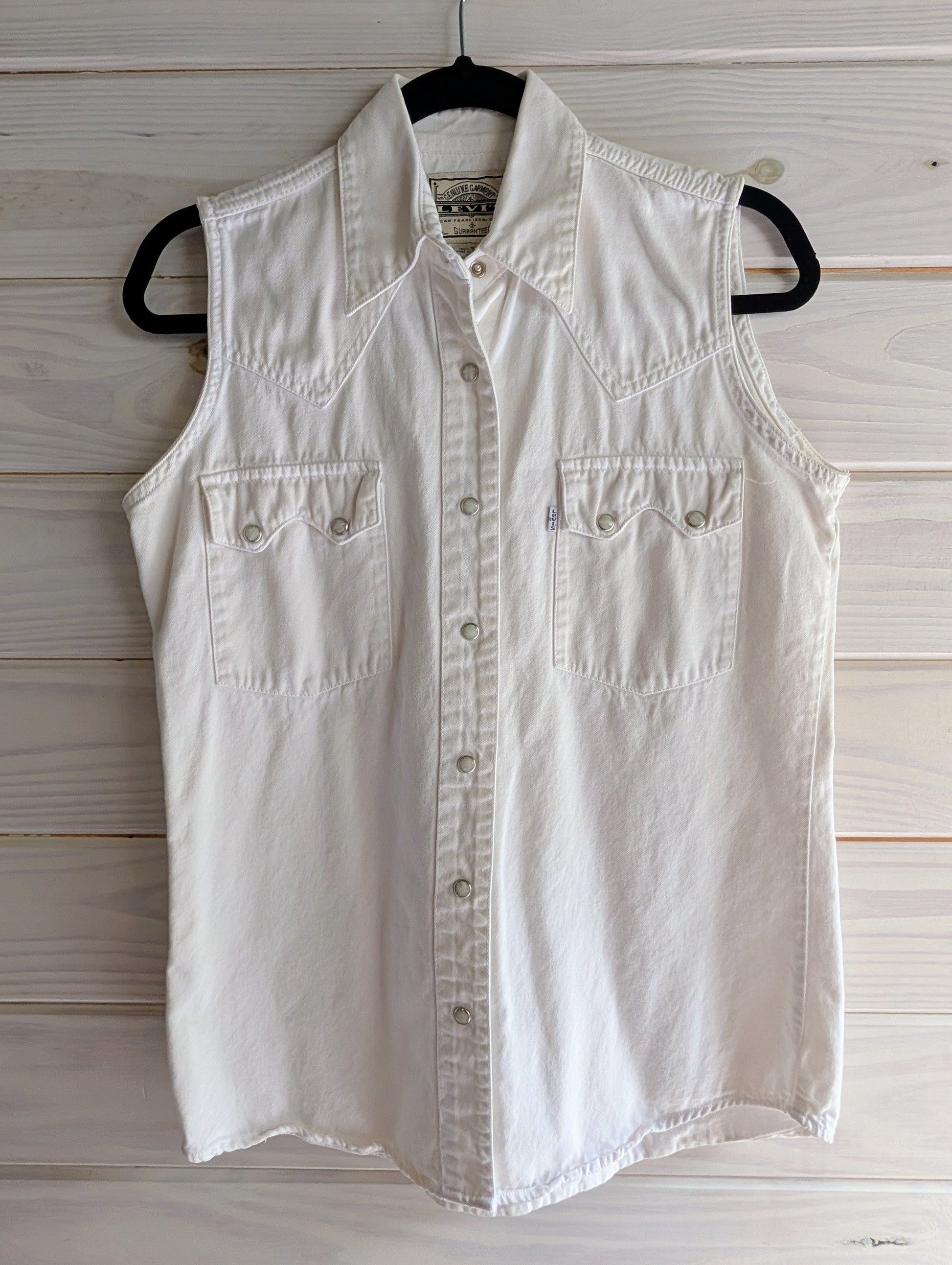 White sleeveless 100% cotton denim shirt with white and silver pearl snap buttons on a hanger against a wooden background.