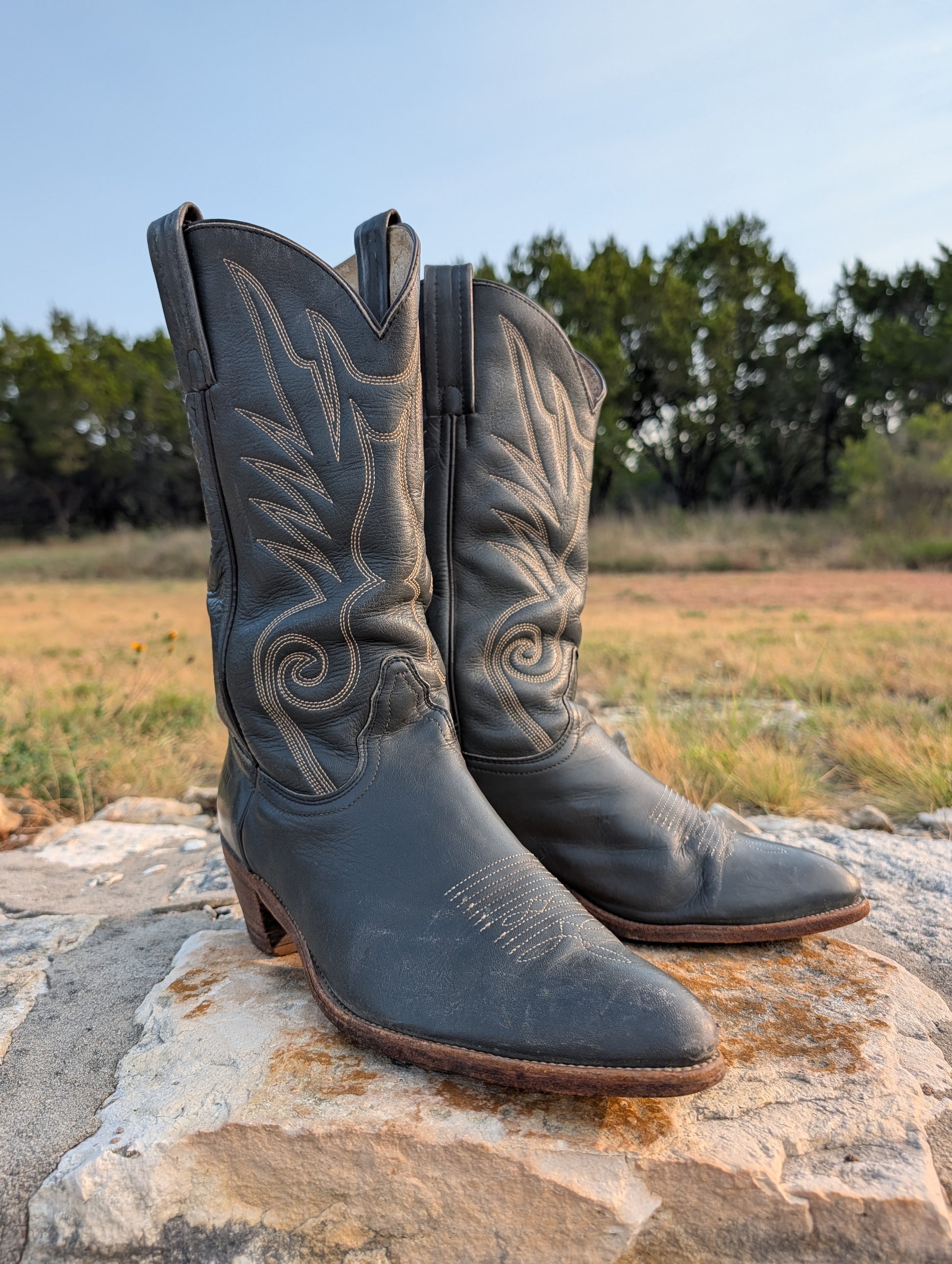 Dark gray leather cowboy boots facing slightly to the right, with decorative stitching in cream thread, a 2" Western heel, resting on a large stone outdoors with a grassy field and trees in the background. 